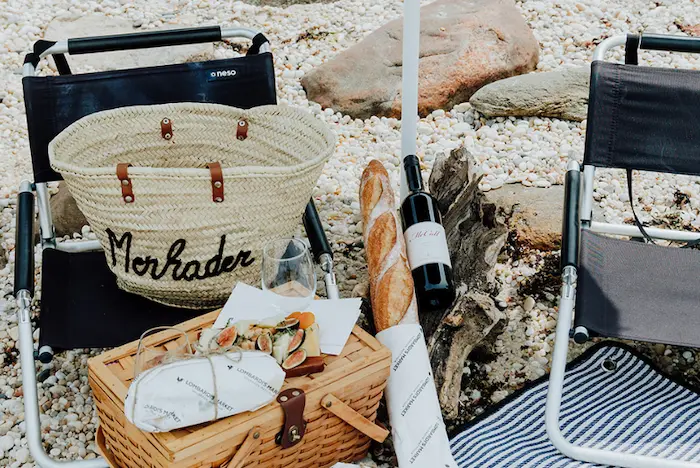 A beach scene with a wicker picnic basket, a bottle of wine, a baguette, and a folding chair.