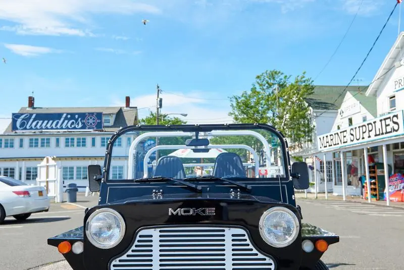 A black Moke vehicle in a parking lot, with a white building and a blue sky in the background.