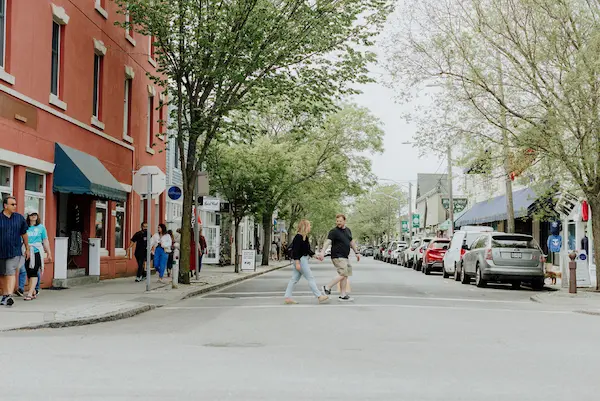 Two people walking on a city street, sidewalk, parked cars, trees, storefronts.