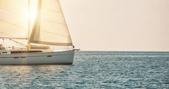 A white sailboat on a large body of water, with the sun shining brightly behind its sails.