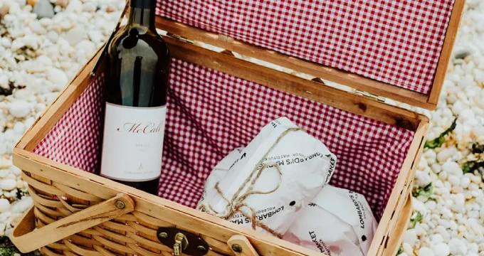 A wooden picnic basket with a red and white checkered lining, containing a bottle of wine and a cloth napkin.