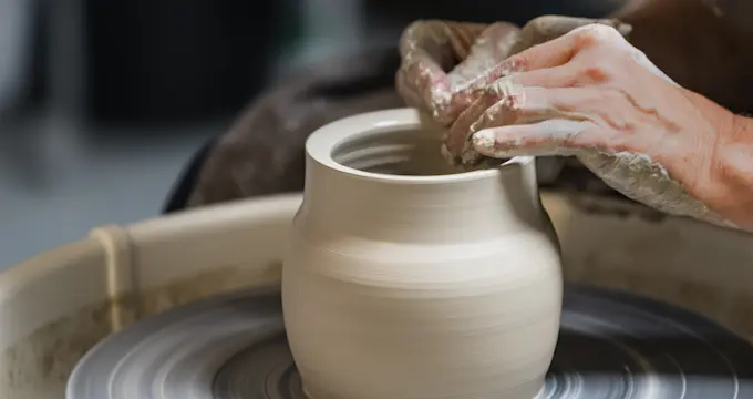 A person's hands shaping a large, white ceramic pot on a pottery wheel.
