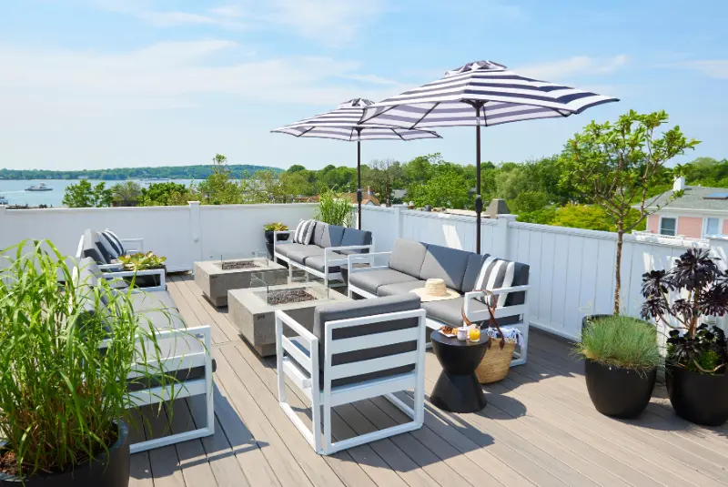A rooftop patio with a striped umbrella, gray furniture, and potted plants. The patio overlooks a body of water, with trees and a blue sky in the background.