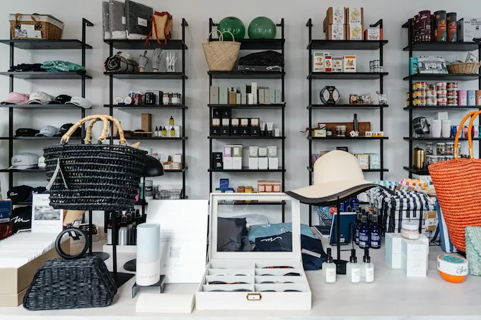 A cluttered room with shelves and various items on display. The shelves are black and hold a mix of products, including bags, hats, and other merchandise. The room appears to be a store or showroom, with a white floor and a white wall in the background.