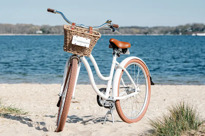 A white bicycle with a wicker basket on the handlebars, parked on a sandy beach with a body of water in the background.