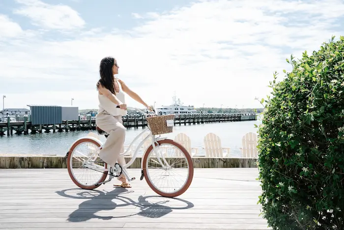 Woman riding a bike on a dock with a bush and body of water in the background.