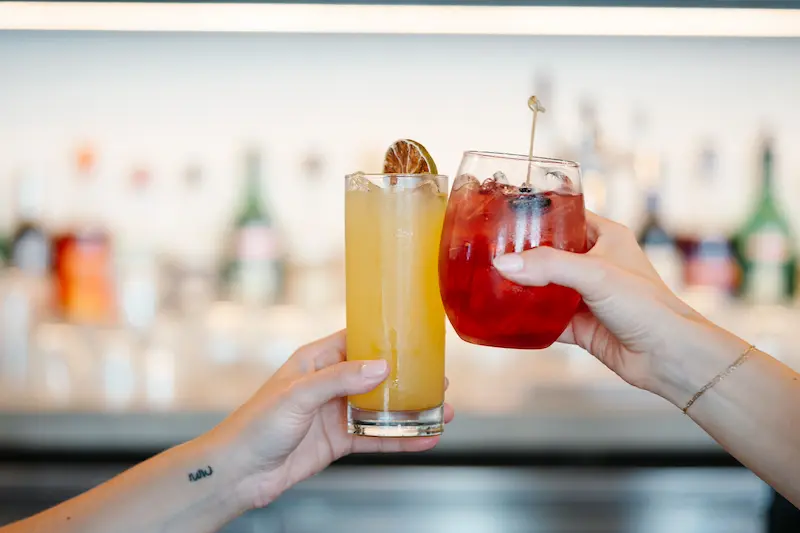 Two hands holding drinks, with a blurred bar in the background.