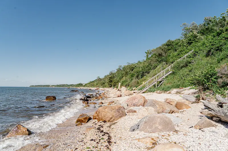 A rocky beach with a wooden walkway and greenery on a sunny day.