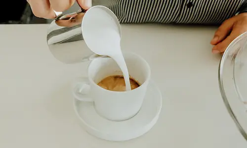 A hand pouring milk into a cup of coffee from a metal pitcher, viewed from directly above.