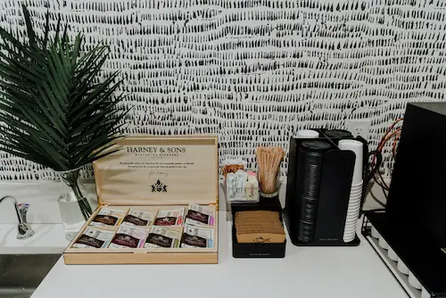 A white countertop with a box of Harney & Sons tea, a coffee maker, and a plant in front of a white and black patterned wall.