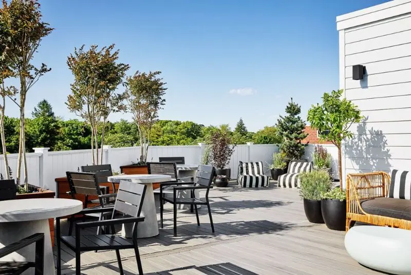 A patio with a table and chairs, a couch, potted plants, and a fence. The patio is surrounded by trees and a blue sky.