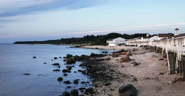 A beach scene with a house, rocks, and water. The house is white with a dark roof. There are rocks in the water and on the shore. The sky is blue with clouds.