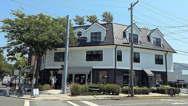A two-story building with a black and white exterior, featuring a black roof and a black awning over the entrance. The building has a row of windows on both floors, and a tree stands in front of it.