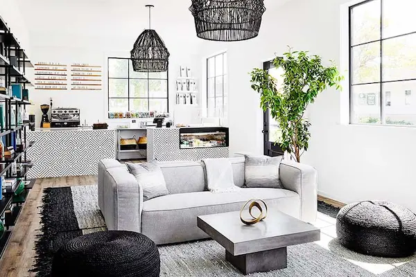 black wicker light fixture, white couch, plant, coffee table, black pouf, bookshelf, window, desk, shelving, bookcase, rug, coffee maker, books, wall art, herringbone rug, white walls, wood floor.